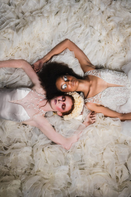 Photograph of two women in wedding dresses lying on the floor on a large white cloth wedding dress fabric with their heads attached.