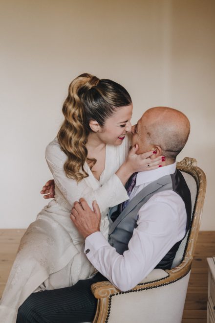 Photograph of the bride and groom in which the groom is seated in an armchair with the bride sitting on his knee and caressing him affectionately.