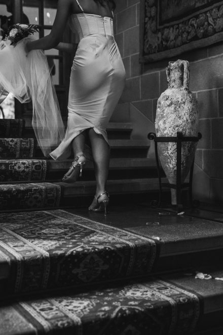 Black and white photograph of the bridesmaid holding the train of the bride's dress as they walk up a flight of stairs.