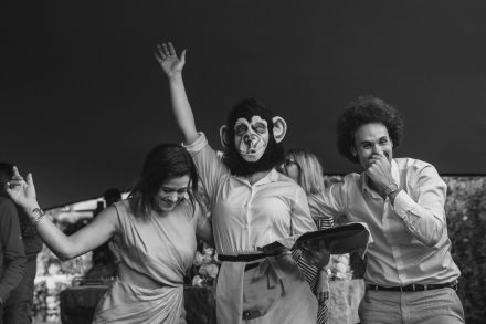Black and white photograph of two guests posing with a catering waitress with a monkey mask on.