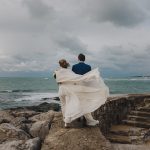 Photograph of the bride and groom walking along the seashore on a day of rough seas.