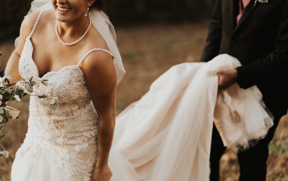 Woman dressed as a bride walking while a man holds the train of her dress so that she does not step on it.