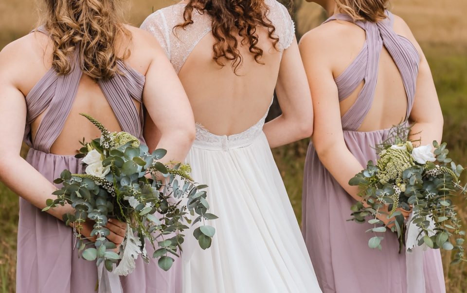 Back photograph of the bride dressed in white with her two bridesmaids at her sides dressed in violet carrying bouquets of flowers.