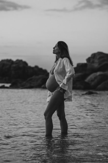 Black and white pregnancy photo of Ale on the seashore with an open shirt exposing her belly.
