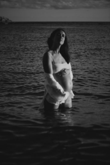 Black and white pregnancy photo of Helena on the seashore with an open shirt exposing her belly.