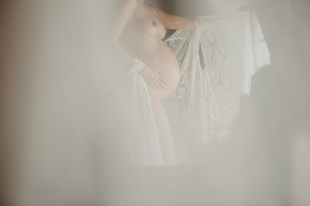Artistic photograph of half-naked Xenia caressing her belly from behind a curtain.